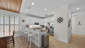 A modern kitchen with white cabinets, a central island with bar stools, stainless steel appliances, and a ship's wheel decoration on the wall. - 4BR Pool Game Room | Good Morning Rentals (Jacksonville Beach)
