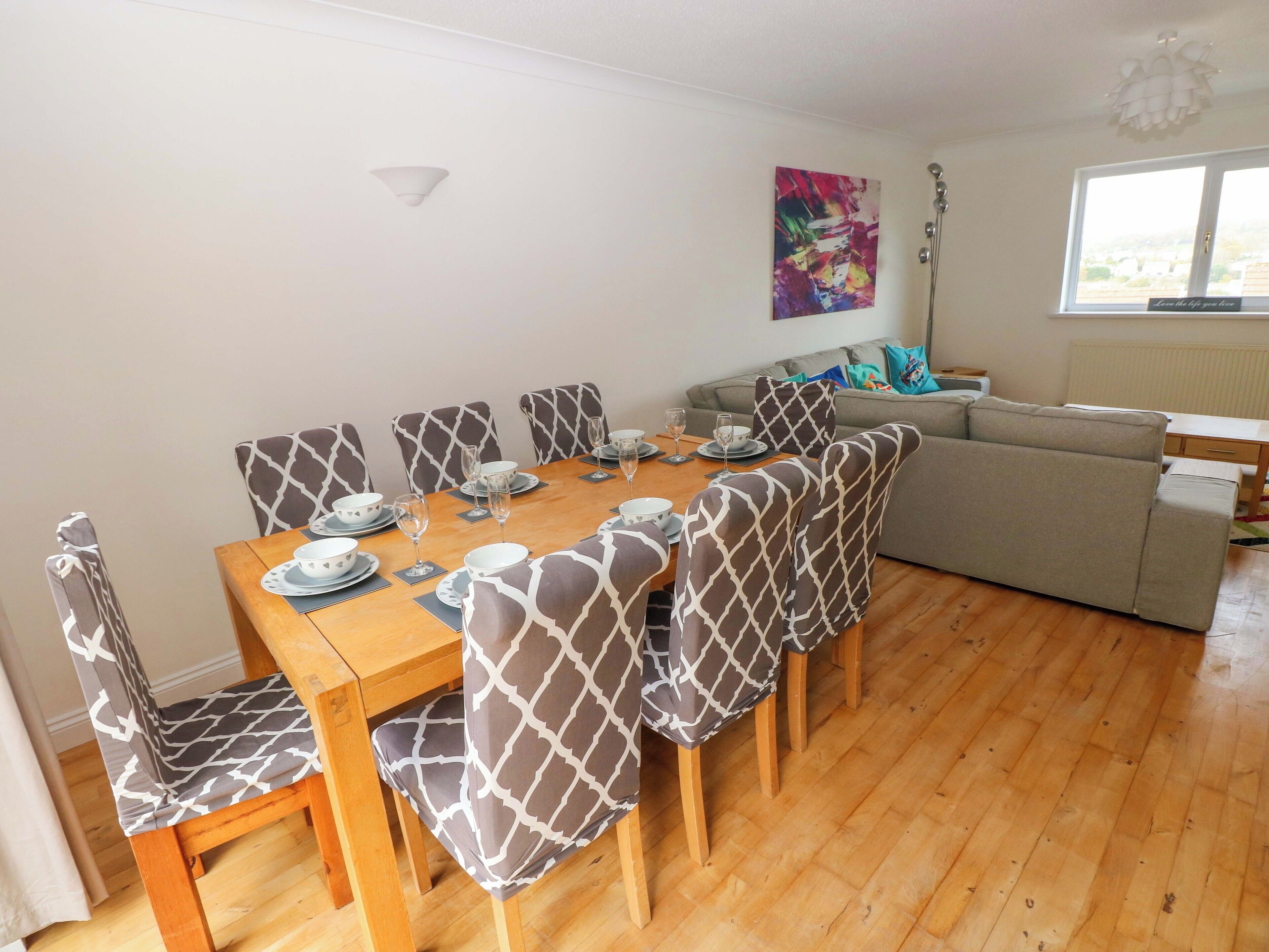 A dining area with a wooden table set for six, surrounded by chairs with a giraffe pattern. There is a sofa and a window in the background.