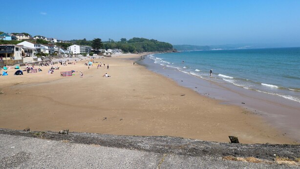 Une vaste plage de sable fin, un ciel bleu dégagé, quelques personnes et des bâtiments en arrière-plan.
