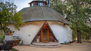 Une maison en forme de dôme géodésique, dotée d’une porte en bois, entourée d’arbres et bordée d’une allée en gravier.