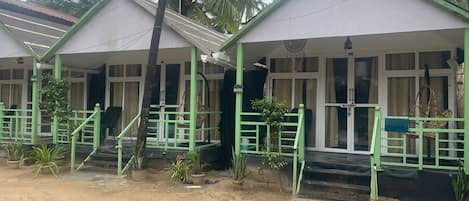 A row of small, single-story buildings with green railings and white walls, surrounded by palm trees.