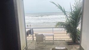 A covered outdoor area with two plastic chairs and a table, overlooking a beach with waves and a palm tree. - Palolem Sea n' Sand Beach View   (Canacona)