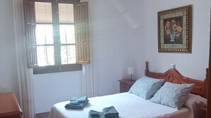 A bedroom with a bed, a wooden dresser, a chair, a window with curtains, a hanging lamp, and a framed picture on the wall. - Esperanza's little house (Aracena)