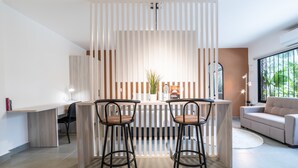 Un salon moderne doté d’un îlot central, de deux tabourets de bar, d’un canapé et d’une grande fenêtre offrant une vue sur la verdure.