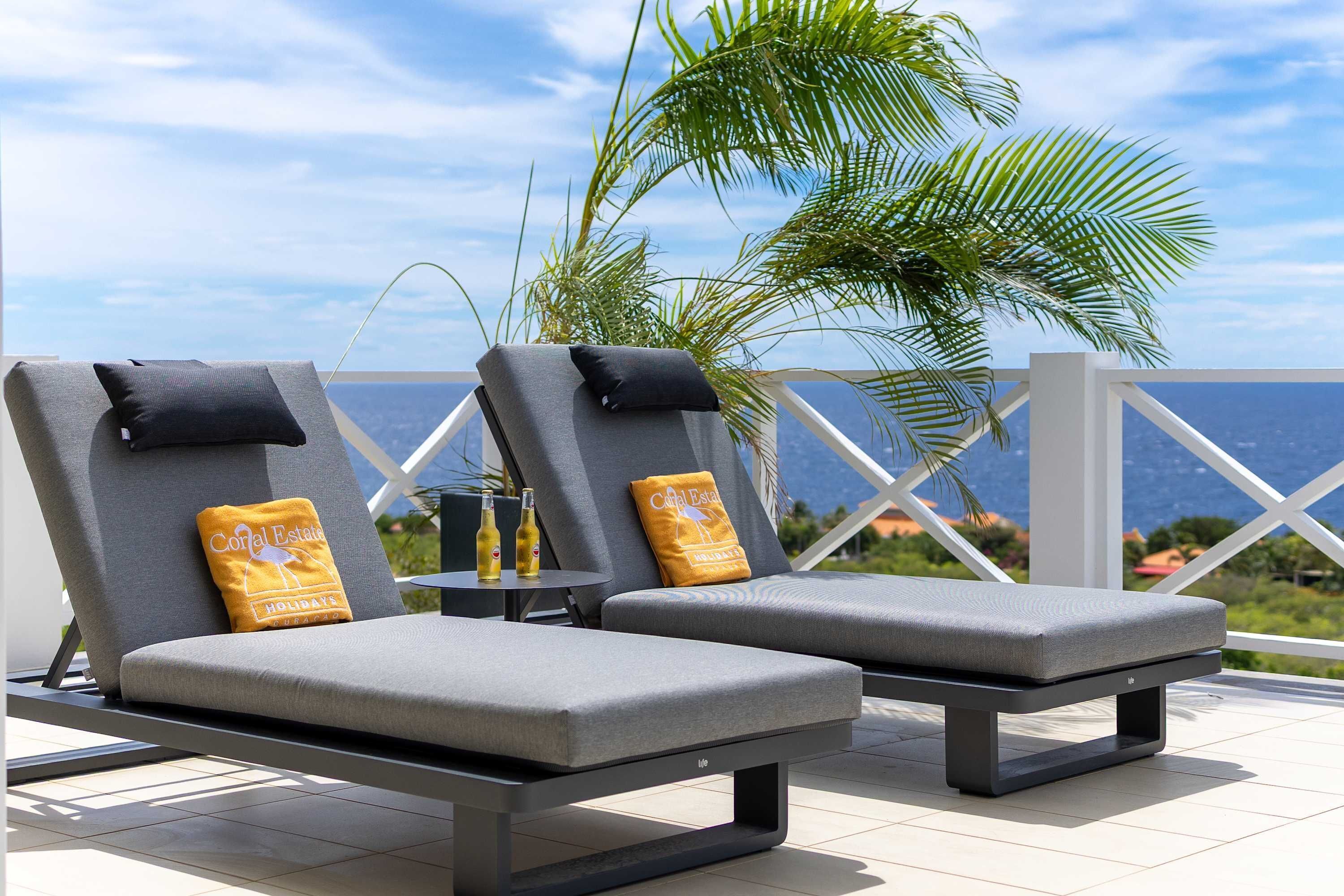 Two lounge chairs with cushions, a small table with bottles, and a view of the ocean.