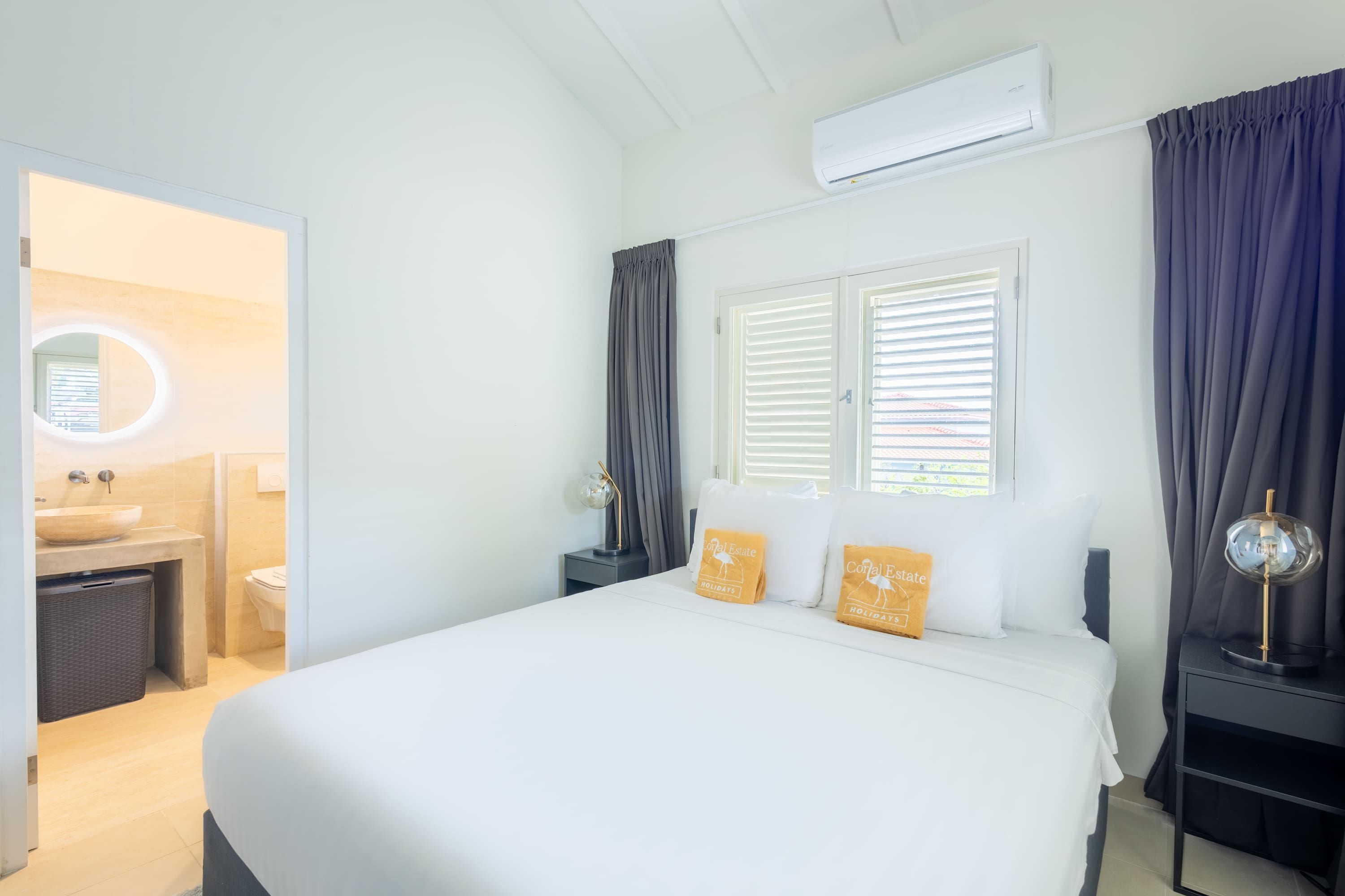 Une chambre d’hôtel avec un grand lit, deux oreillers, une table de chevet, une commode, un miroir, une fenêtre avec des stores et un appareil de climatisation.