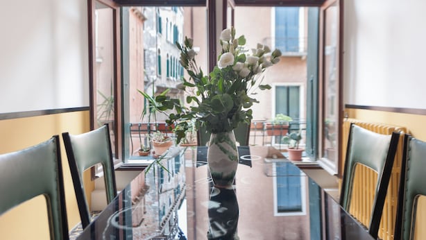 Ett matbord med stolar, en vas med blommor och en utsikt över byggnader genom öppna fönster.