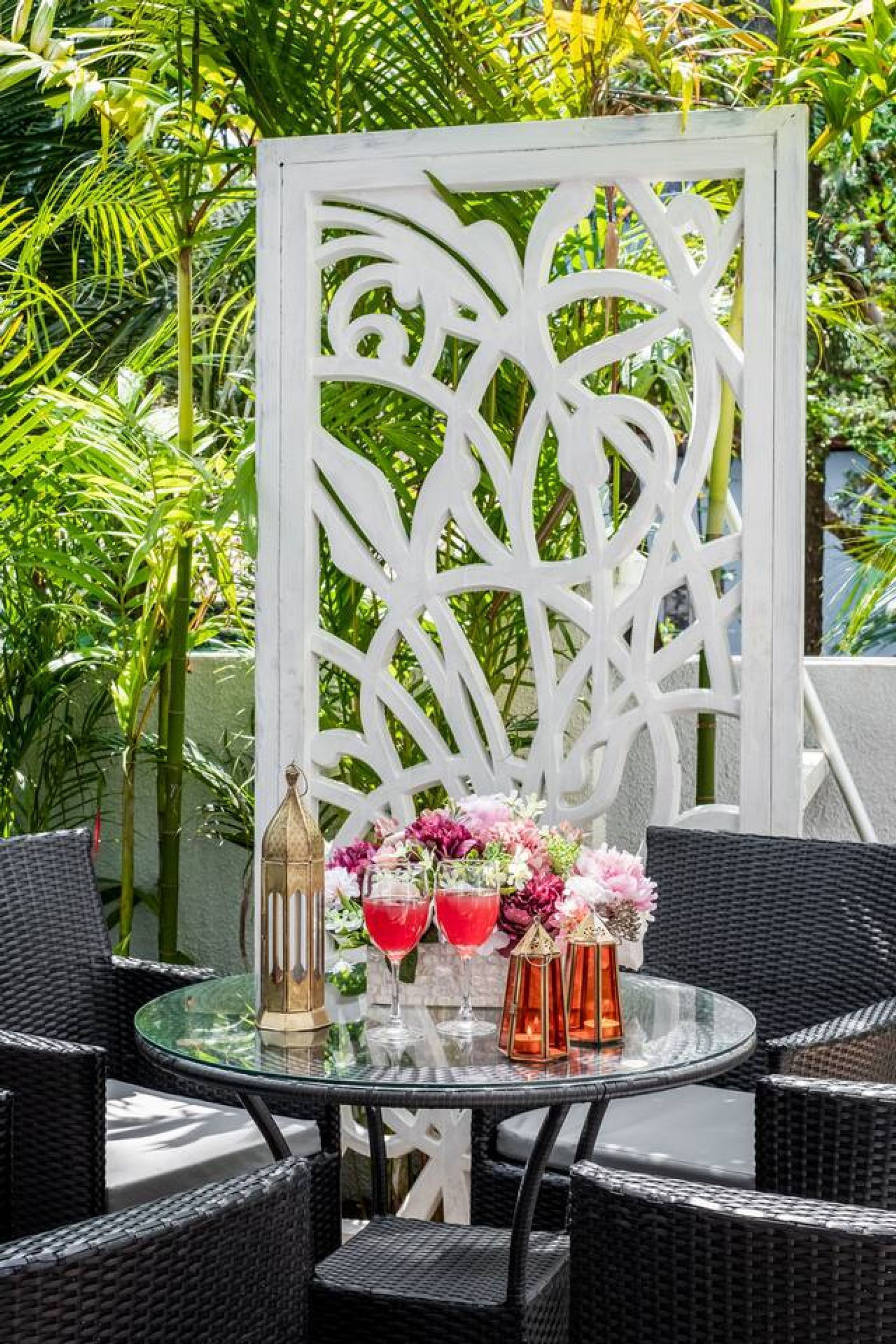A round glass table with wicker chairs, a decorative white partition, and a vase with pink flowers and red drinks.