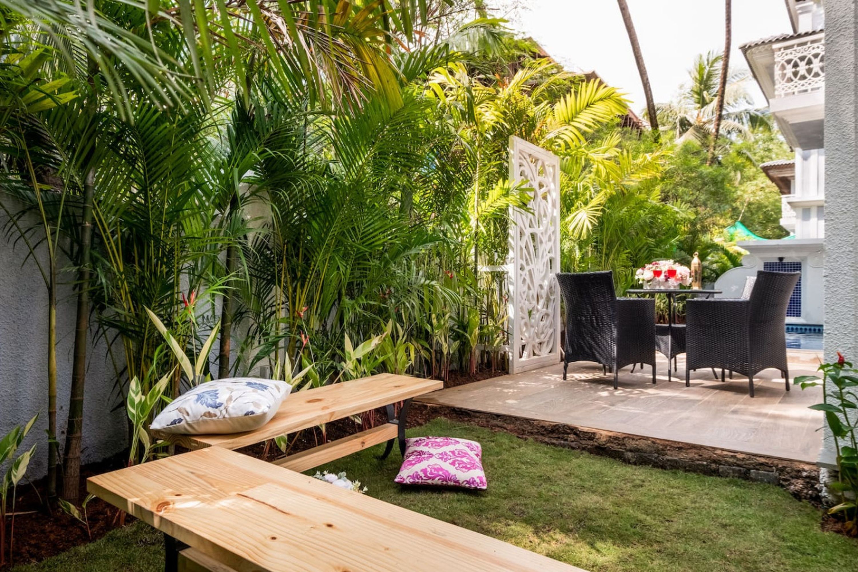 Une terrasse avec une table et des chaises en bois, entourée d’une végétation luxuriante et d’une clôture blanche décorative.