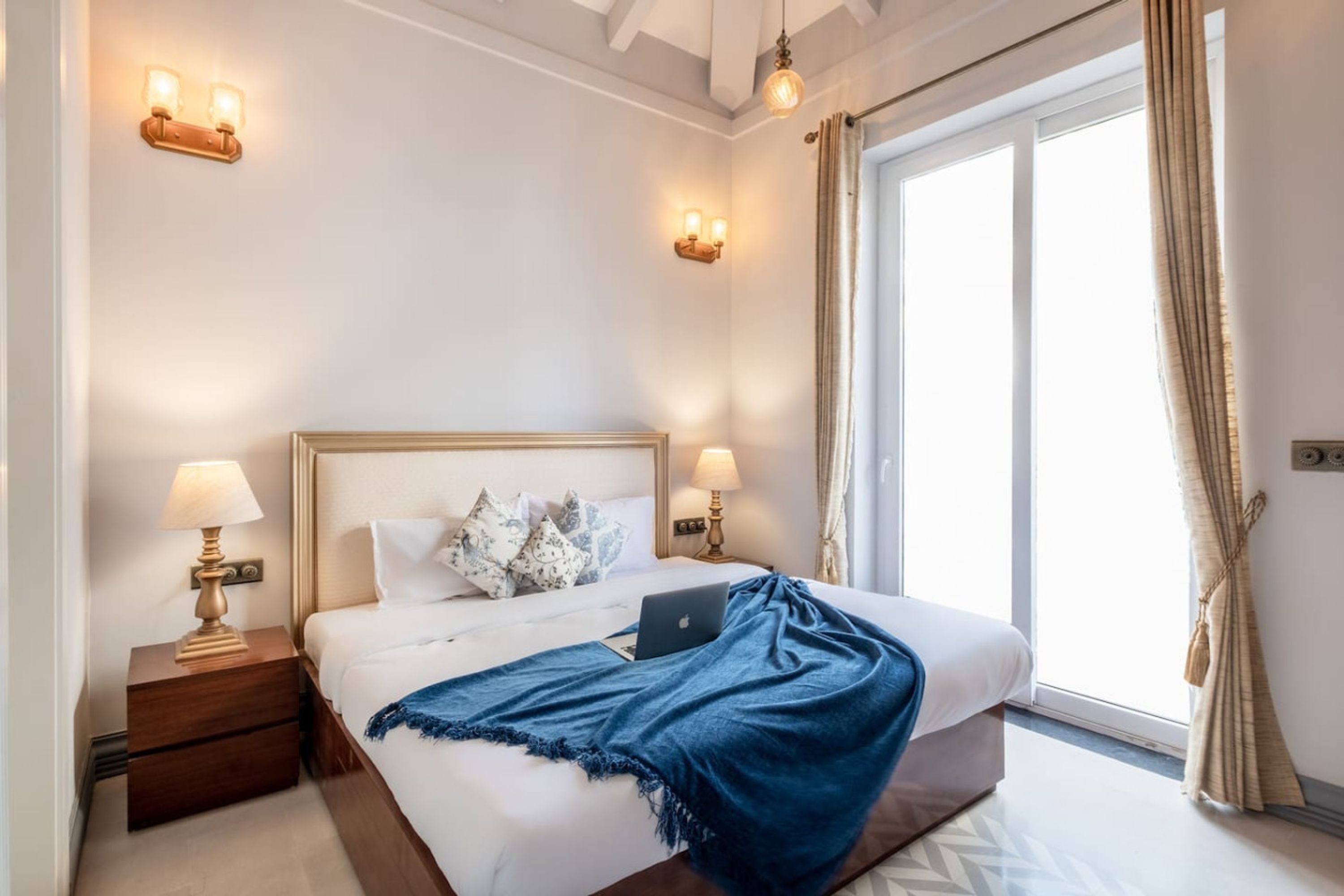 Hotellihuone, jossa on suuri sänky, kaksi yöpöytälamppua, kannettava tietokone sängyllä ja ikkuna, jossa on verhot.