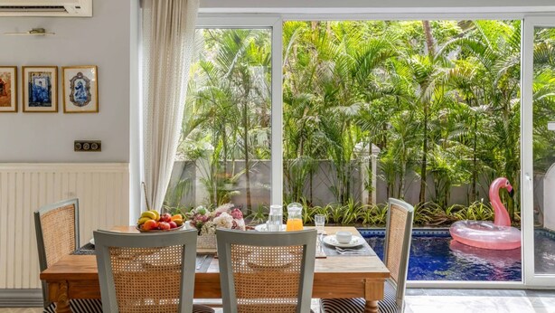 A dining area with a table set for two, a pink flamingo float in a pool, and a view of lush greenery through large windows. - Luxury 4bhk Pool Getaway in North Goa Villa Kaia (Valpoy)