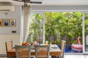 A dining area with a table set for two, a pink flamingo float in a pool, and a view of lush greenery through large windows. - Luxury 4bhk Pool Getaway in North Goa Villa Kaia (Valpoy)
