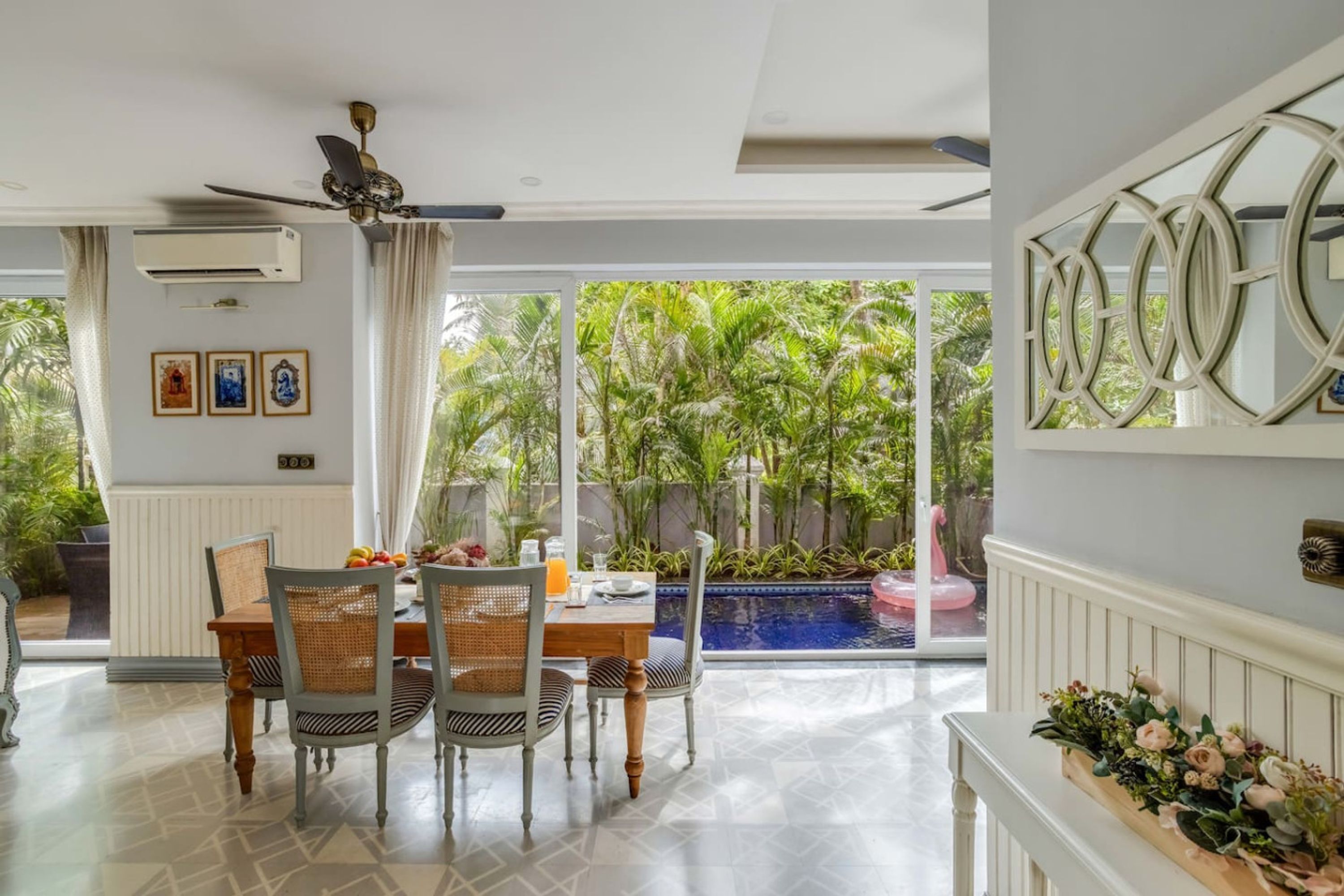 Zona pranzo con tavolo e sedie, ventilatore a soffitto e vista su piscina e vegetazione.