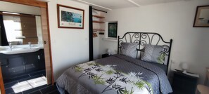 Une chambre à coucher comprenant un lit, une table de chevet, une commode, un miroir et une salle de bain avec un lavabo, ainsi qu’un tableau encadré accroché au mur.