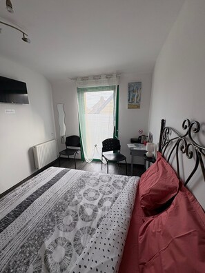 Ein Schlafzimmer mit Bett, Schreibtisch, Stuhl, Heizkörper, Fernseher und einem Fenster mit Vorhang.