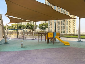 A playground with a slide, swings, and a climbing structure, surrounded by a shaded area and a view of high-rise buildings. - Creekside 18 A - 103 2BD (Dubai)