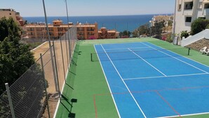 A tennis court with a net, surrounded by a chain-link fence, and overlooking a coastal area with buildings and a clear sky. - La Casa Bonita (Benalmádena)
