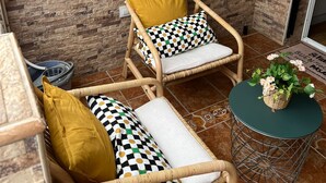 A wicker chair with a white cushion and a mustard-colored pillow, a small round table with a woven basket, and a wall with a brick pattern and string lights. - La Casa Bonita (Benalmádena)