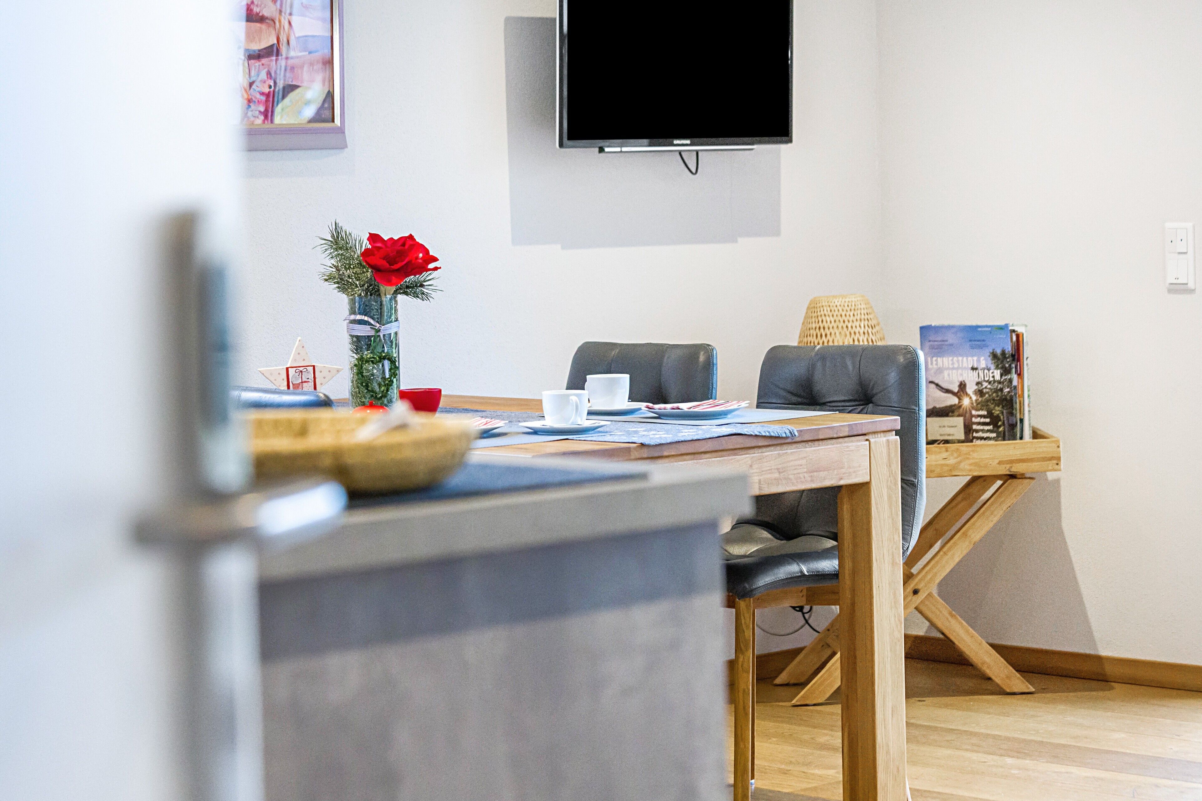 A modern living room with a flat-screen TV, a dining table with chairs, a coffee table, and a decorative vase with a red flower.