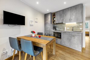 Une cuisine moderne avec des meubles gris, une table à manger en bois avec des chaises, un téléviseur à écran plat fixé au mur et une petite fenêtre.