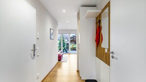 A hallway with wooden flooring, white walls, a door with a mirror, and a view to an outdoor area with a red chair.