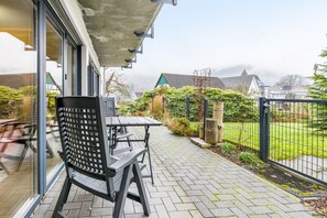 An outdoor patio with a table and chairs, a glass door, and a view of a garden and houses in the background. - Apartment 'Buchfink B, 50 sqm' with Mountain View, Private Terrace and Wi-Fi (Lennestadt- Saalhausen)