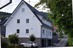 Une maison blanche à deux étages, avec un toit en pente, une voiture garée devant et un arbre au premier plan.