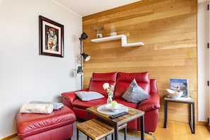Un canapé en cuir rouge, une petite table en bois avec une télécommande, un tableau encadré accroché au mur et une étagère blanche ornée d’objets décoratifs.