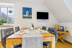 A dining area with a table set for four, a TV mounted on the wall, and a window with a view of trees.