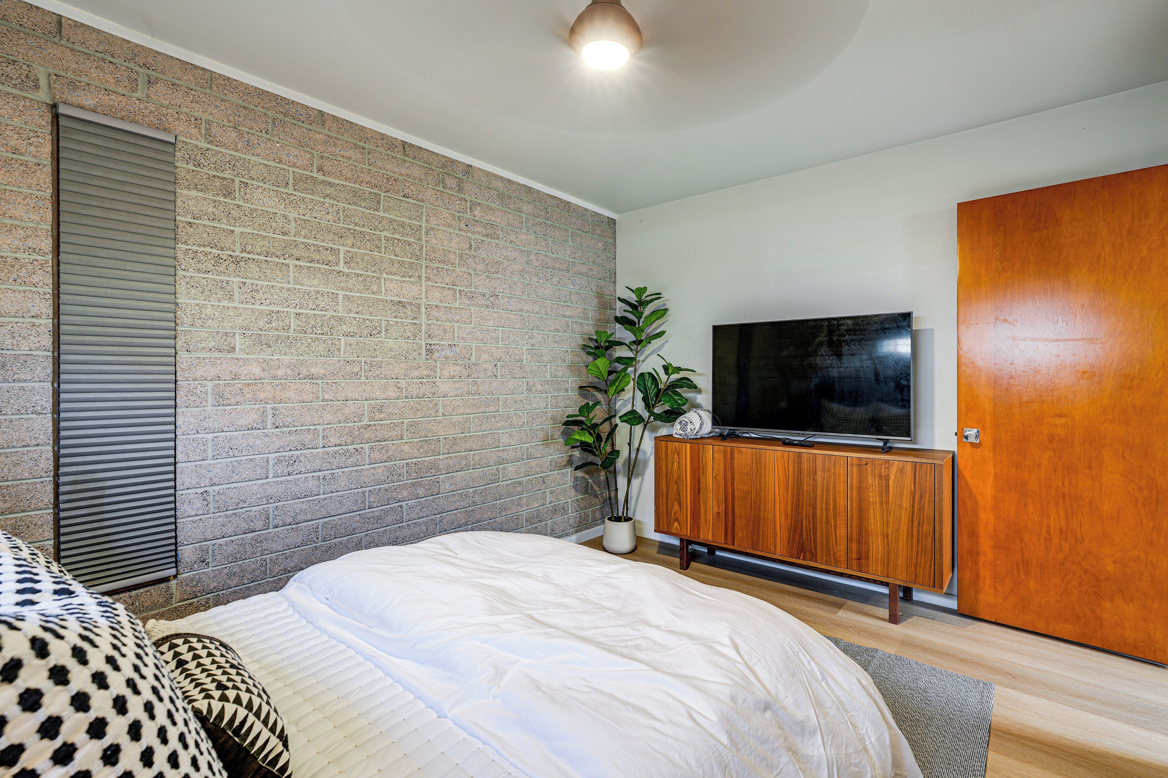 A bedroom with a brick wall, a bed with a white duvet, a wooden cabinet with a TV, and a wooden door.