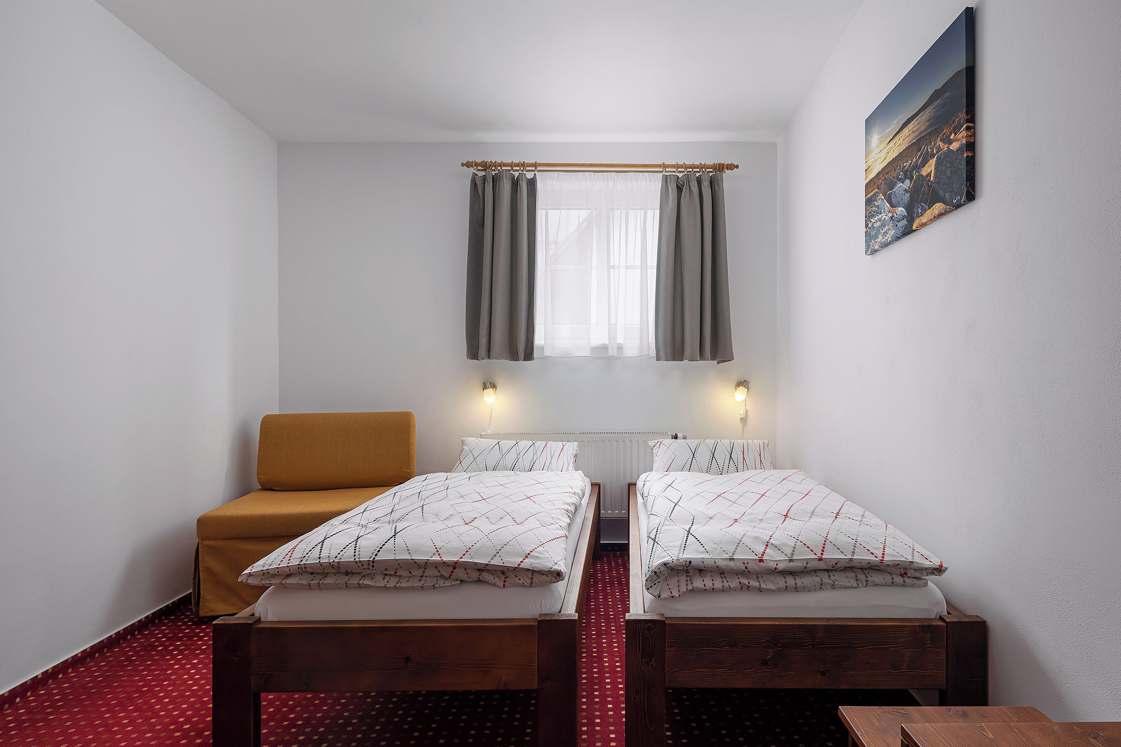 A small, clean room with two single beds, a wooden bench, a red patterned carpet, and a window with curtains.