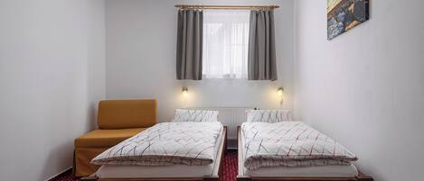 A small, clean room with two single beds, a wooden bench, a red patterned carpet, and a window with curtains.