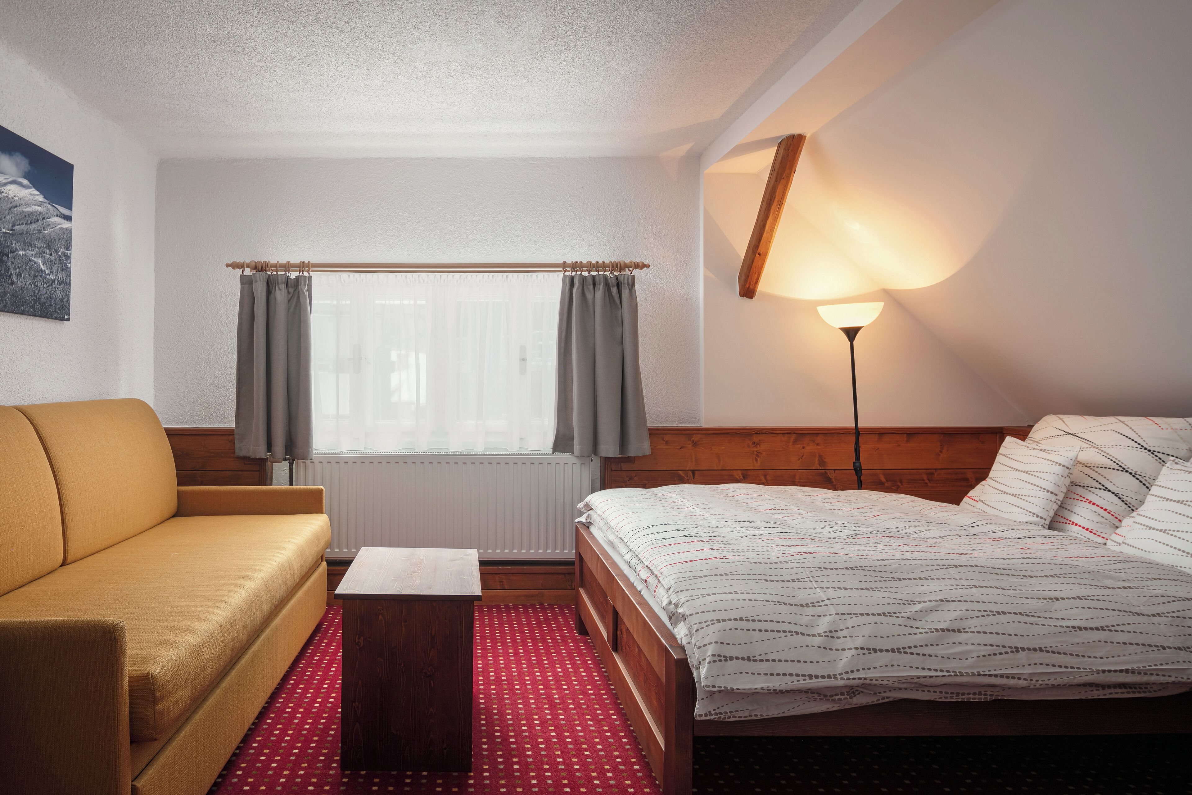 A hotel room with a wooden bed, a yellow sofa, a small table, and a window with curtains.