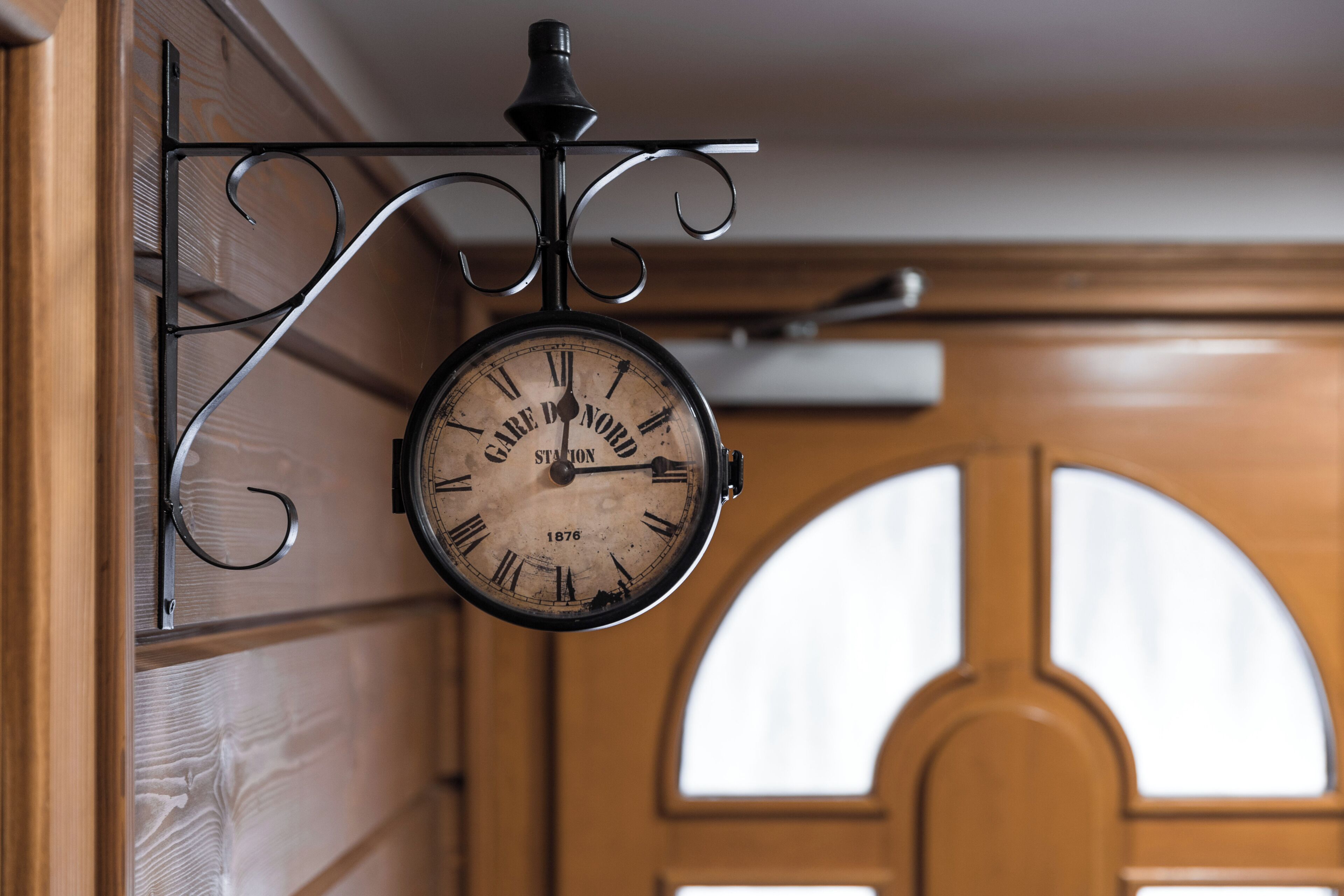 A vintage wall clock with Roman numerals and the year 1876, mounted on a black metal bracket.