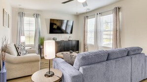 Un salon avec un canapé gris, un fauteuil beige, une table basse ronde, un lampadaire, un téléviseur fixé au mur et un ventilateur de plafond.