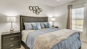 Une chambre à coucher comprenant un lit, des tables de chevet, un ventilateur de plafond, une œuvre d’art murale et une fenêtre avec des rideaux.