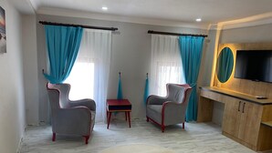 A modern bedroom with two armchairs, a small table, a TV, and a large window with curtains. - FETHİYE OLD TOWN SUİTES (Fethiye)