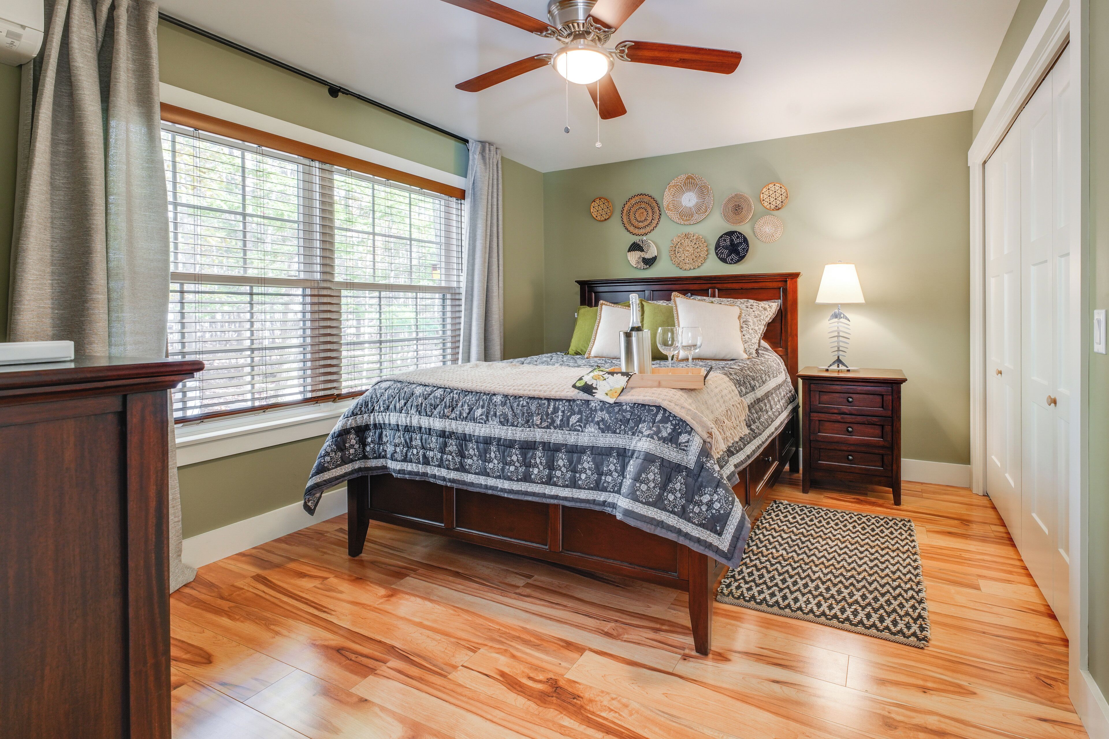 Une chambre avec un plancher en bois, un lit recouvert d’une couvre-lit à motifs, un ventilateur de plafond, une table de chevet avec une lampe et une fenêtre avec des stores.