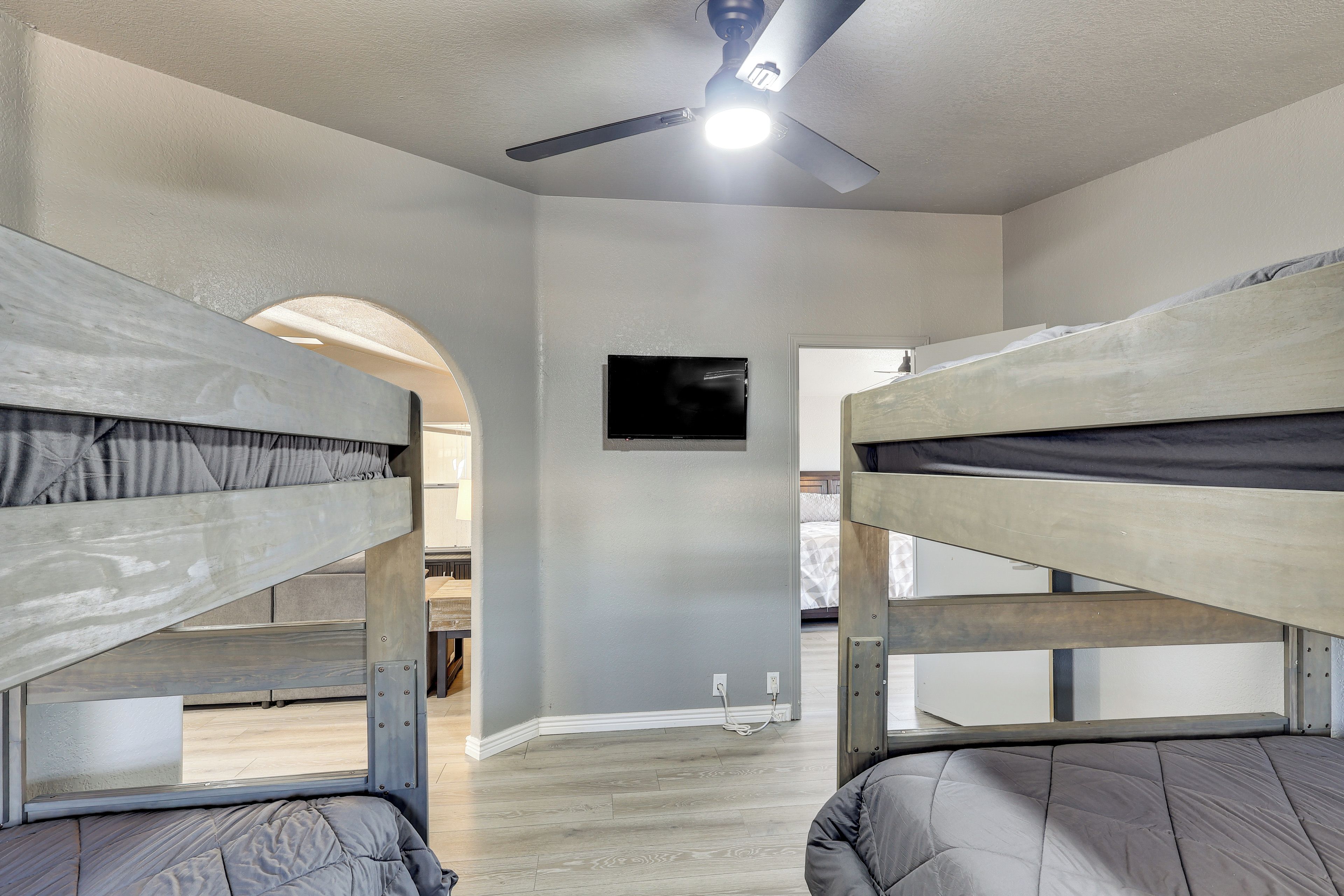 A room with bunk beds, a ceiling fan, a TV mounted on the wall, and a wooden floor.