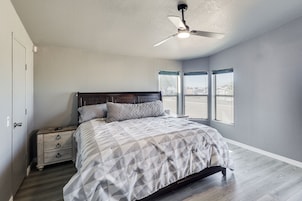 Un dormitorio con cama, mesitas de noche, ventilador de techo y ventanas amplias.