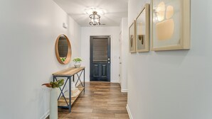A hallway with a wooden floor, a console table with a plant, a mirror, and framed artwork on the wall.