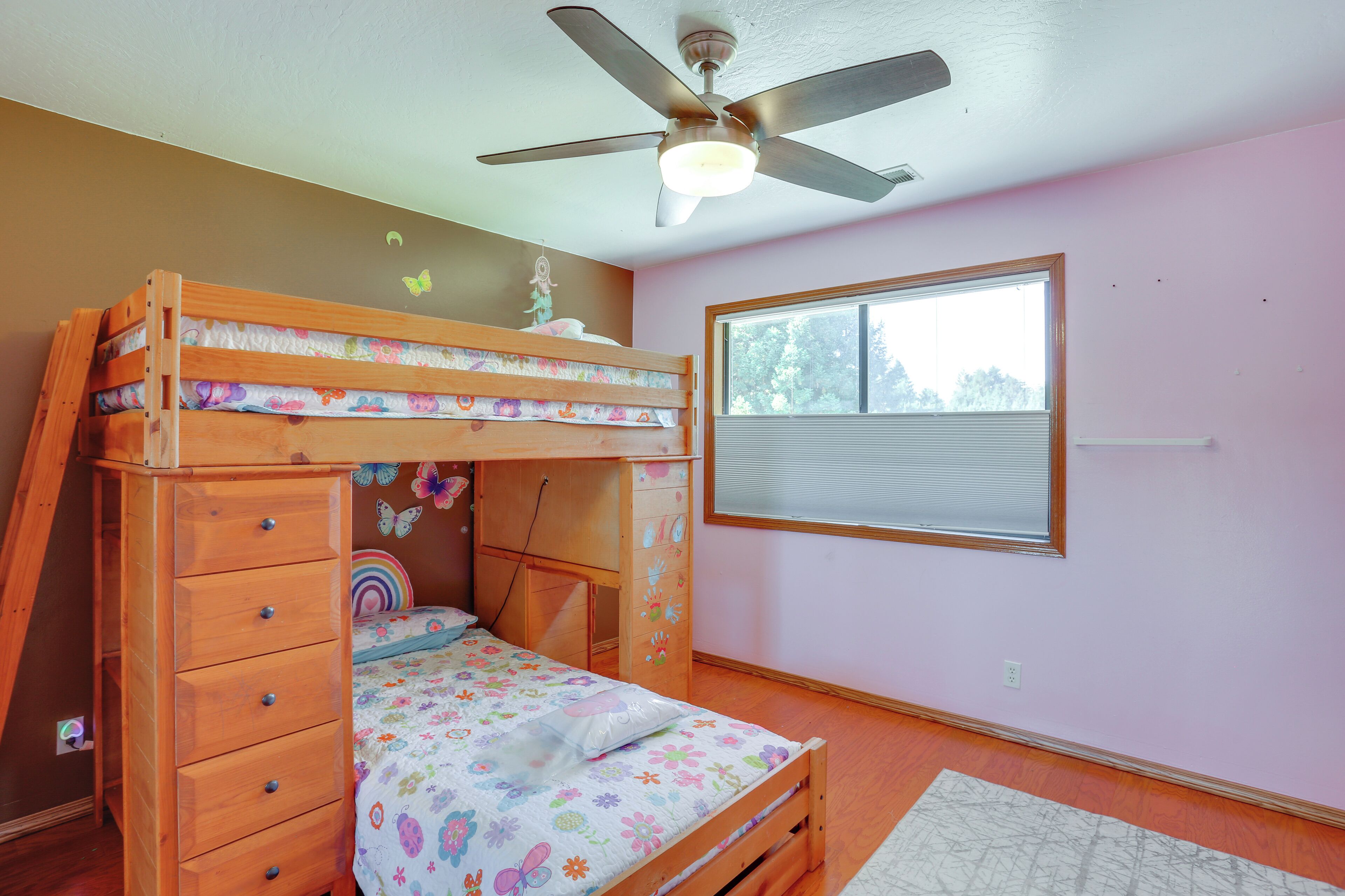A bunk bed with a wooden frame, a ceiling fan, a window with blinds, and a wall with butterfly decals.