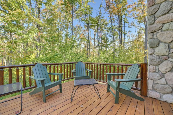 A wooden deck with two Adirondack chairs and a table, overlooking a forested area.