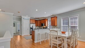 A spacious kitchen with wooden cabinets, a central island, and a dining area with white chairs. - Stay Kureous - Just 3 blocks from the beach with Rooftop Views & Multiple Outdoo (Kure Beach)