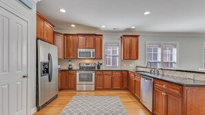 A kitchen with wooden cabinets, stainless steel appliances, and a patterned rug. - Stay Kureous - Just 3 blocks from the beach with Rooftop Views & Multiple Outdoo (Kure Beach)