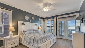 A bedroom with a bed, nightstands, a ceiling fan, and a view through a door. - Stay Kureous - Just 3 blocks from the beach with Rooftop Views & Multiple Outdoo (Kure Beach)
