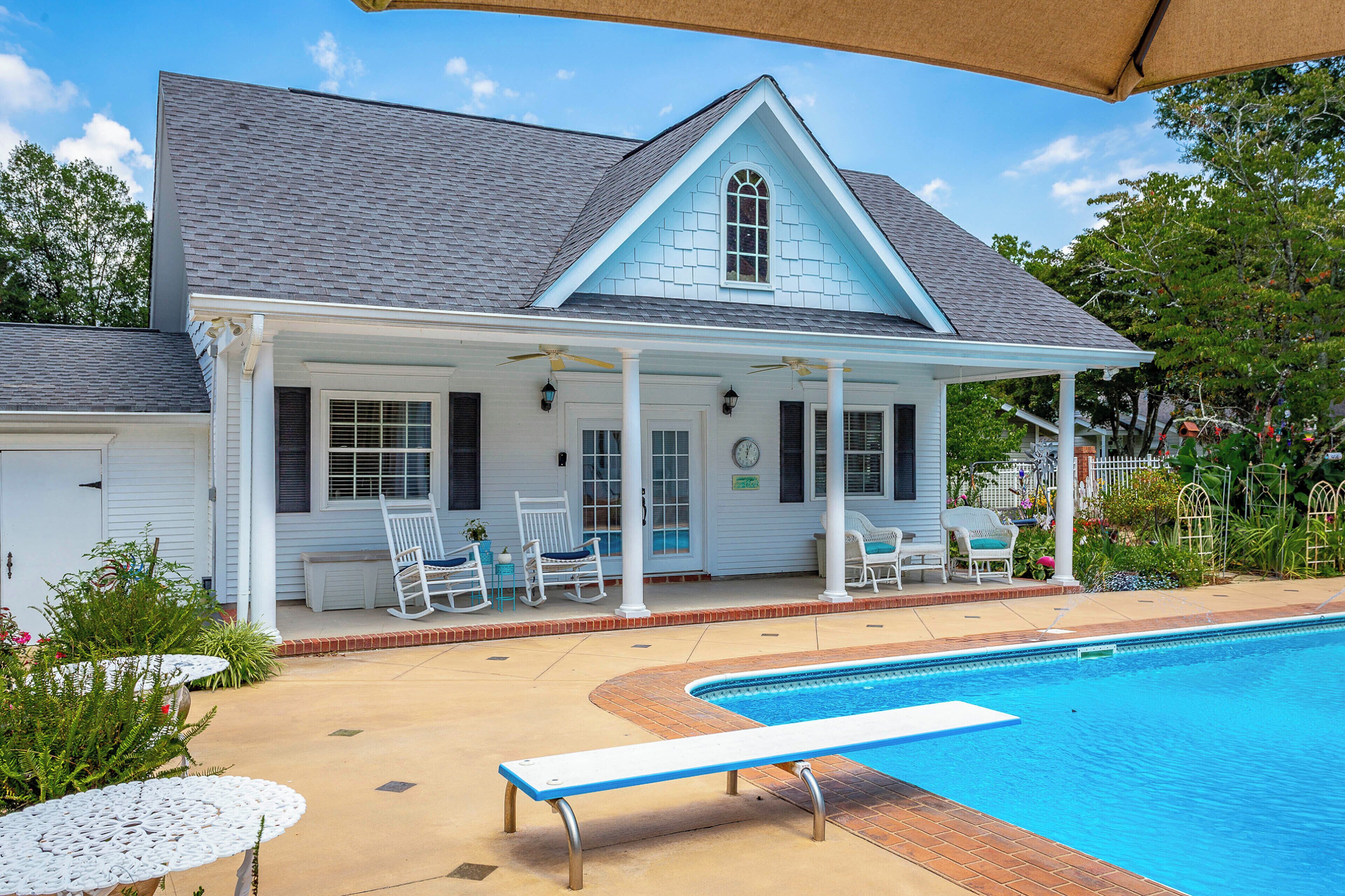 A house with a pool, white exterior, and a covered porch with chairs.