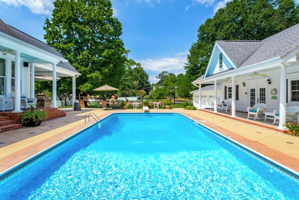 A rectangular swimming pool with a clear blue water surface, surrounded by a brick deck and white lounge chairs.