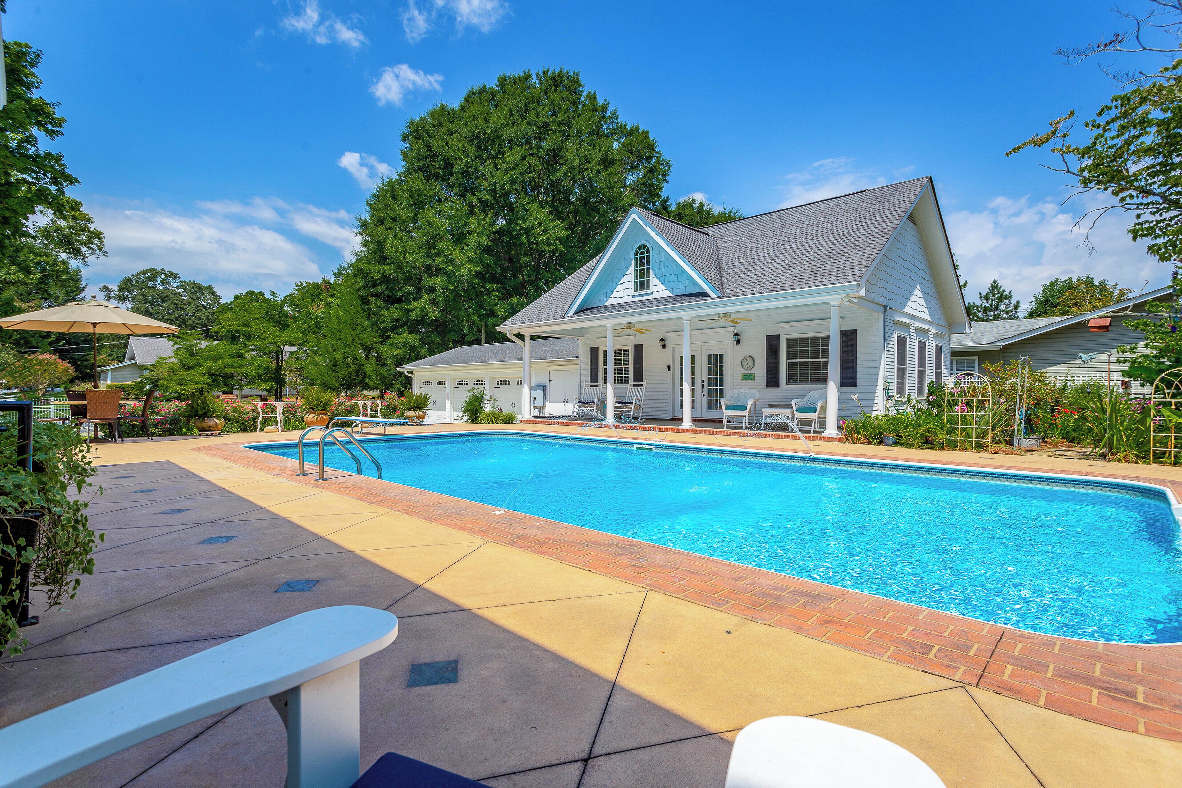 A swimming pool with a covered patio area, a white building with a blue roof, and a large tree in the background.