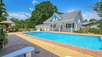 A swimming pool with a covered patio area, a white building with a blue roof, and a large tree in the background.